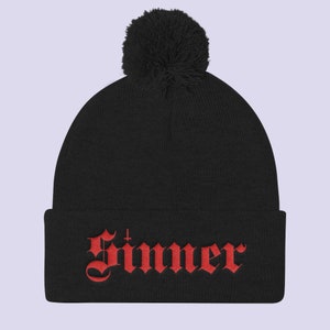 May include: Black beanie with a red embroidered "Sinner" with a cross above the "n". The beanie has a black pom pom on top.