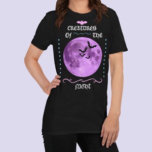 以下が含まれることがあります： 黒いTシャツに紫色の月とコウモリが描かれており、ピンク色の文字で「Creatures of the Night」と書かれています。