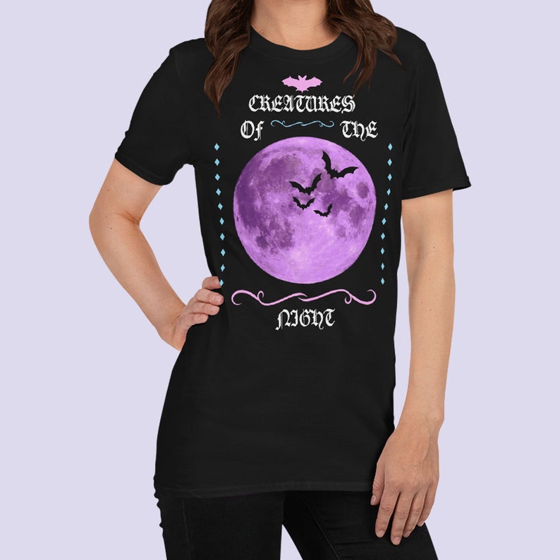 以下が含まれることがあります： 黒いTシャツに紫色の月とコウモリが描かれており、ピンク色の文字で「Creatures of the Night」と書かれています。