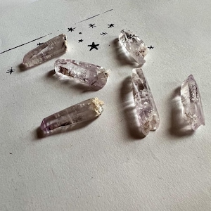 May include: Six amethyst crystal points with varying sizes and shapes are arranged on a white surface. The crystals have a pale purple hue with some areas appearing translucent. A simple black line drawing of stars and a shooting star is in the background.