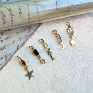 Puede incluir: Cinco dijes de metal dorado con varias formas de piedras preciosas y motivos colgantes. Los dijes incluyen una piedra marquesa roja con una estrella, una piedra ovalada negra con una lágrima, una piedra marquesa clara con una llave, una piedra marquesa morada con una luna creciente y una piedra ovalada clara con un sol.