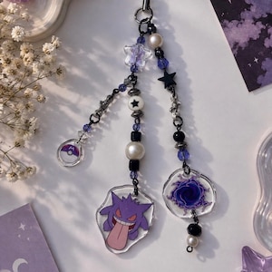 May include: A phone charm featuring three dangling strands of beads, including white pearls, black spheres, purple crystals, and star-shaped charms. The charms include a purple Gengar character, a swirling purple portal, and a small circular Poké Ball icon.
