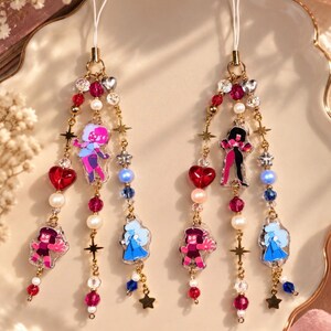 Op de afbeelding: Twee bijpassende telefoonhangers met goudkleurige kettinkjes, versierd met rode hartvormige kralen, witte parels, heldere kristallen en kleine gouden sterretjes. Elke hanger bevat drie acryl figuurtjes in rood, blauw en roze.