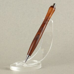 May include: A wooden pen with a brown and dark brown wood grain pattern. The pen is sitting in a clear plastic stand.