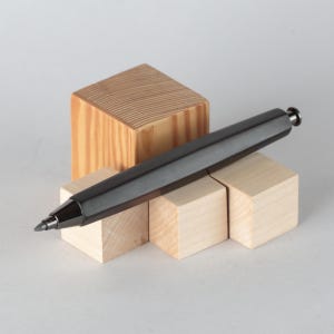 Puede incluir: Un portaminas gris oscuro descansa sobre pequeños bloques de madera. El lápiz tiene un diseño elegante y moderno con un acabado metálico. Los bloques de madera son de color claro y de forma cuadrada, con un bloque más grande en el fondo.