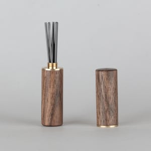 Puede incluir: Un difusor de aceites esenciales de madera con bandas doradas. El difusor contiene cañas negras. La tapa está separada. La veta de la madera es visible. El difusor mide unos 10 cm de alto.