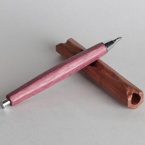Puede incluir: Un bolígrafo artesanal con un vibrante cuerpo de madera rosa y morado y una punta plateada. El bolígrafo descansa sobre un portalápices de madera marrón. El bolígrafo es un instrumento de escritura elegante.