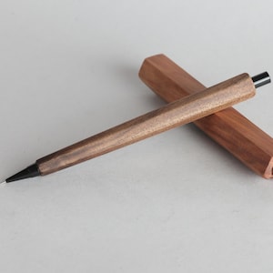Op de afbeelding: Een houten pen met een zeshoekige schacht en een zwarte punt, naast de bijpassende houten etui. De pen en etui zijn gemaakt van warmbruin hout en de pen heeft een strak, minimalistisch ontwerp. De pen is ongeveer 15 cm lang.