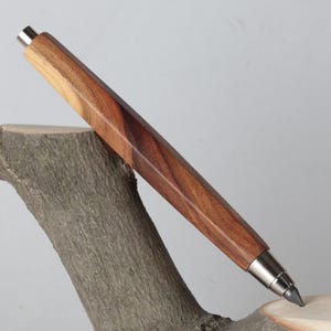 Puede incluir: Un portaminas de madera con diseño geométrico. El cuerpo del lápiz es de madera de color marrón oscuro con detalles metálicos plateados. Descansa sobre un trozo de madera.