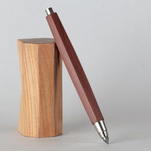 May include: A dark brown wooden mechanical pencil with silver accents leans against a light brown wooden block. The pencil has a geometric design and a silver tip. The block has a unique, twisted shape.