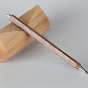 Puede incluir: Un portaminas de madera con acabado marrón oscuro y detalles plateados descansa sobre un bloque de madera marrón claro. El lápiz tiene un diseño elegante y minimalista, perfecto para escribir o dibujar.