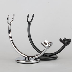 May include: Two shaving brush stands. One is chrome-plated, with a circular base and a curved arm ending in a U-shaped holder. The other is black, with a similar design. Both stands are designed to hold a shaving brush.