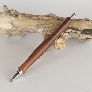 Op de afbeelding: Een handgemaakte houten pen met een donkerbruine afwerking en zilverkleurige metalen accenten. De pen wordt gepresenteerd op een stuk drijfhout, dat de natuurlijke houtnerf en het elegante ontwerp laat zien. Een stijlvol schrijfinstrument.