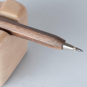 Puede incluir: Un portaminas de madera marrón oscuro con punta de metal plateado, apoyado en un soporte de madera clara. El lápiz tiene forma hexagonal y una superficie lisa y pulida. Ideal para escribir y dibujar.