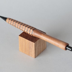 May include: A wooden mechanical pencil with a geometric design on the grip, resting on a small wooden block. The pencil has a black tip and eraser, and is made of light brown wood.
