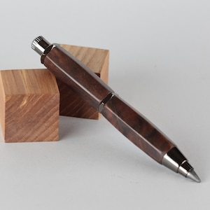 Puede incluir: Un portaminas de madera marrón oscuro con un diseño geométrico. El lápiz tiene una punta y detalles plateados. Descansa sobre dos pequeños bloques de madera marrón claro.