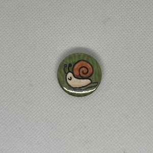 Puede incluir: Un botón verde con un caracol de dibujos animados en él. El caracol es marrón y blanco con una hoja verde detrás.