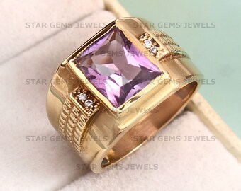 May include: A gold-colored ring with a large, square, light purple gemstone at the center. The ring has a thick band with decorative detailing and small accent stones on each side of the main stone. The ring is displayed in a white box.