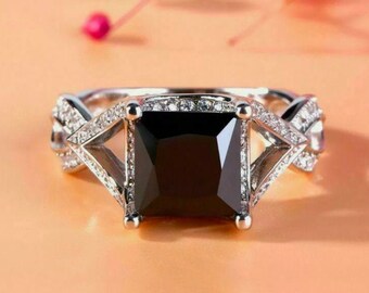 May include: A silver ring with a large black square-shaped gemstone surrounded by smaller clear gemstones. The ring has a delicate, intricate design with a twisted band.