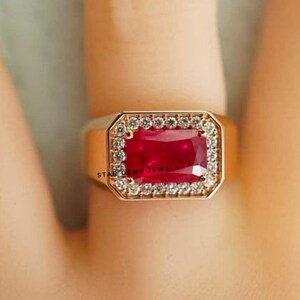 Könnte beinhalten: Ein goldener Ring mit einem großen, rechteckigen, roten Rubin-Edelstein, der von einem Heiligenschein aus kleineren, weißen Diamanten umgeben ist. Der Ring ist in einer goldenen Band gefasst und hat die Gravur "STAR GEMS JEWELS" an der Seite.