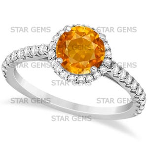 Op de afbeelding: Een zilveren ring met een grote, ronde, oranje edelsteen omringd door kleinere, heldere edelstenen. De ringband is ook bezet met kleine, heldere edelstenen. De tekst "STAR GEMS" is zichtbaar.
