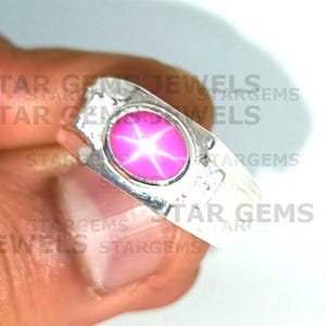 May include: A silver ring featuring a large, oval, pink star sapphire gemstone. The gemstone has a prominent six-ray star pattern. The ring is held by a person.