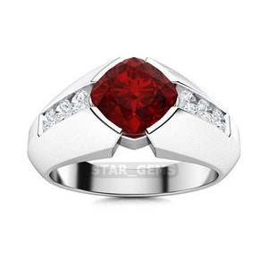 May include: A men's ring featuring a large, square-cut red gemstone surrounded by smaller white gemstones. The ring is crafted from white metal and has a wide band.