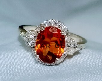 May include: A silver ring with an oval-shaped orange gemstone surrounded by a halo of smaller white gemstones. The ring has a delicate design with additional white gemstones on the band.