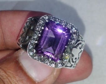 May include: A silver ring with an ornate design featuring a large, rectangular, purple amethyst gemstone surrounded by smaller white gemstones.