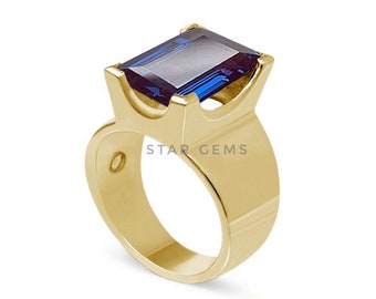 May include: A gold ring with a large, dark blue, emerald-cut gemstone. The ring has a unique, modern design with a wide band and a setting that elevates the gemstone.