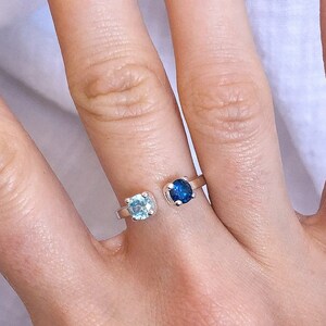 Könnte beinhalten: Ein silberner Ring mit zwei Edelsteinen, einem hellblauen und einem dunkelblauen, an einer Hand. Der Ring hat ein offenes Design, wobei sich die Edelsteine auf beiden Seiten des Fingers befinden. Die Hand liegt auf einer weißen Oberfläche.