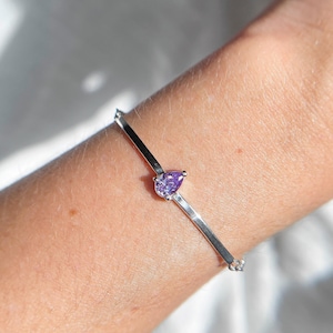 Op de afbeelding: Een zilveren armband met een peervormige, paarse edelsteen. De armband heeft een minimalistisch ontwerp met een dunne, rechte band en een enkele, prominente edelsteen. De armband wordt om een pols gedragen.