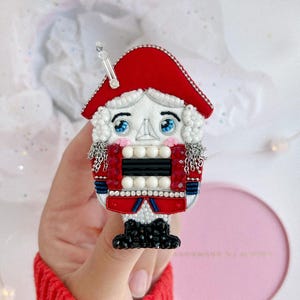 Può includere: Una spilla a forma di schiaccianoci realizzata con perline, caratterizzata da un cappello rosso con un dettaglio argentato, capelli bianchi, occhi azzurri e guance rosa. L'uniforme è decorata con perline bianche e nere, cristalli rossi e fili.