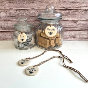 May include: Four glass jars with ceramic labels for tea, sugar, coffee, and biscuits. Each label features a bee design and the word for the item in black text.