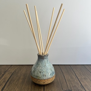 May include: A ceramic reed diffuser featuring a bulbous base with a speckled light blue and white glaze over a textured, unglazed tan bottom. Several thin, straight wooden reeds extend upwards from the narrow neck of the vessel.