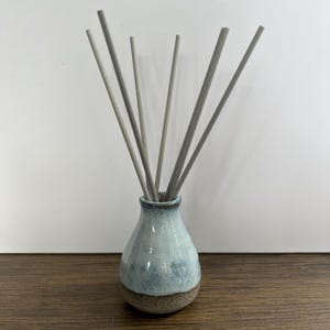 May include: A ceramic reed diffuser with a bulbous base featuring a light blue and white glazed top section and a textured brown bottom. Seven thin, straight, light grey reeds are inserted into the narrow neck of the vessel, fanning outward.