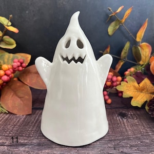 May include: A white ceramic ghost ornament with a smiling face and outstretched arms. The ghost has two eye holes and a jagged mouth. The ornament is a Halloween decoration.