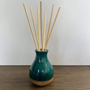 May include: A teal ceramic reed diffuser with a rounded base and a narrow neck. The vessel features a glossy, deep teal glaze on the upper portion and a natural, unglazed tan clay finish at the bottom. Seven light-coloured wooden reeds are inserted.