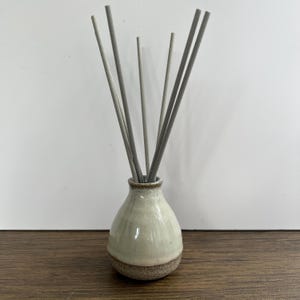 May include: A ceramic reed diffuser featuring a bulbous, cream-coloured base with a speckled, textured brown bottom. Six slender, light grey diffuser reeds are inserted into the narrow neck of the vessel, extending upwards in a fan-like arrangement.