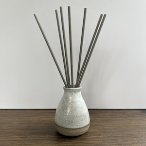 May include: A ceramic reed diffuser featuring a speckled white and cream glaze on the upper portion and a natural brown clay base. Eight thin, straight, grey diffuser sticks are inserted into the narrow neck of the small, rounded vessel.