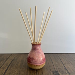 May include: A ceramic reed diffuser featuring a bulbous base with a glossy pink and magenta glaze over a speckled tan bottom. Eight thin, light-coloured wooden reeds are inserted into the narrow neck, fanning outward in a radial arrangement.