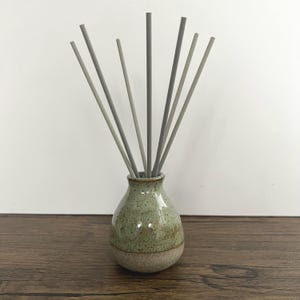 May include: A small, speckled green ceramic vase with a light brown base holds several grey diffuser sticks. The vase sits on a dark wooden surface against a plain white background. The vase has a glossy finish.