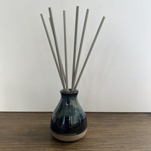 May include: A small, bulbous ceramic vase with a glossy dark blue and black glaze on the upper portion and a textured, light beige base. Seven thin, straight, light grey reed diffuser sticks are inserted into the narrow opening of the vase.