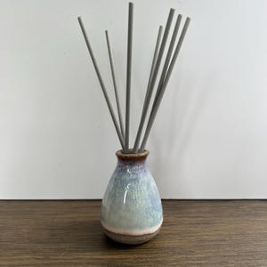 May include: A ceramic reed diffuser with a bulbous base featuring a gradient glaze of light blue, white, and purple tones. The neck of the vessel is a dark brown, and it contains seven thin, straight, light grey reeds extending upward from the opening.