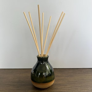May include: A ceramic reed diffuser featuring a rounded, bulbous base with a glossy dark green glaze on the upper portion and a natural, unglazed tan finish on the bottom. Several thin, light-coloured wooden reeds extend upward from the narrow neck.