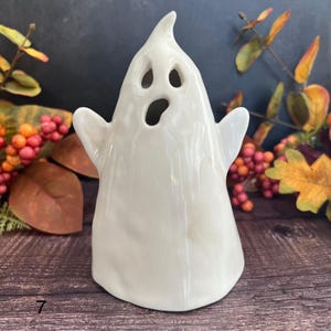 May include: A white ceramic ghost figurine with cut-out eyes and mouth, and outstretched arms. The ghost has a glossy finish and is designed for Halloween decor. The figurine is approximately 13 cm tall.