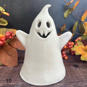 May include: A white ceramic ghost figurine with a smiling face, open arms, and a curved top. The number "10" is printed in black at the bottom left. The background includes autumn foliage.