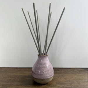 May include: A ceramic reed diffuser featuring a small, bulbous vase with a speckled light pink glaze on the upper portion and a textured, unglazed tan base. Seven slender, straight grey reeds are inserted into the narrow opening of the vase.