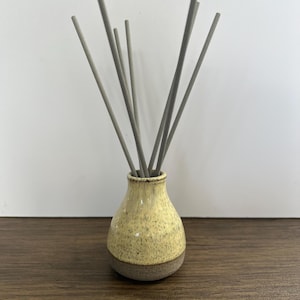 May include: A ceramic reed diffuser featuring a bulbous, speckled pale yellow body with a dark grey base. Several thin, straight grey reeds extend upward from the narrow opening at the top of the vessel, creating a fan-like arrangement.