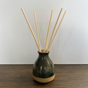 May include: A ceramic reed diffuser featuring a bulbous base with a dark green and blue glazed upper section and a speckled beige lower section. Five light-coloured wooden reeds are inserted into the narrow neck of the vessel, extending upwards.
